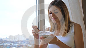 Woman Eating Cereal With Milk Out Of Bowl Standing At Window And Looking Outside