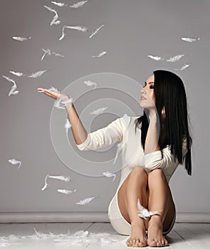 Young pretty brunette woman in mini dress sitting on floor and catching white feathers meaning softness after laser hair