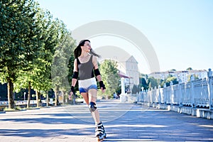 Young pretty brunette woman, riding roller blades in city park in the morning. Fit sporty girl, wearing black top and white shorts