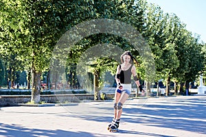 Young pretty brunette woman, riding roller blades in city park in the morning. Fit sporty girl, wearing black top and white shorts