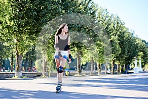 Young pretty brunette woman, riding roller blades in city park in the morning. Fit sporty girl, wearing black top and white shorts