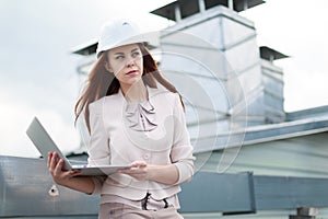 Young, pretty businesswoman in beige suit, brown trousers and helmet sit on the roof and hold laptop