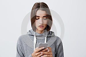 Young pretty caucasian girl with dark long hair in gray hoodie holding smartphone in hands, looking throw social media