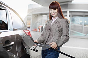 Young pretty Caucasian red haired woman in trendy casual outfit, refilling petrol in her luxury modern car at self