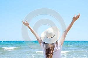 Young Pretty Caucasian Woman with Long Chestnut Hair in Hat Hands Lifted up in the Air Stands at Beach Looks at Turquoise Sea