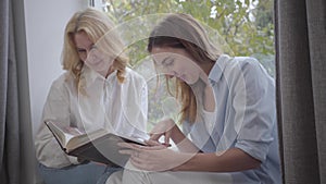 Young pretty Caucasian woman looking photos with older blond lady. Mother and daughter sharing memories at home. Happy