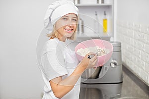 Young pretty Caucasian woman, professional confectioner in white uniform and chef& x27;s hat, standing in the light kitchen