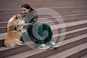 A young pretty Caucasian woman is sitting on the steps in the city center with her little red shepherd dog. Welsh corgi