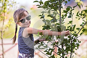 Young pretty child girl in fashionable sun glasses hugging green small tree in summer outdoors