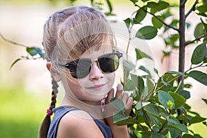 Young pretty child girl in fashionable sun glasses hugging green small tree in summer outdoors