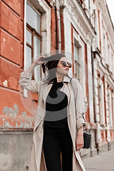 Young pretty cool beautiful woman model in trendy sunglasses in fashionable beige trench coat walks near vintage building in city