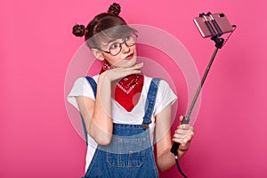 Young pretty dark haired women taking selfie on cell phone, using selfie stick, keeps hands under chin, dressed denim overalls and