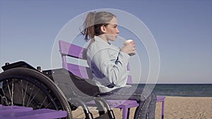 Young Pretty disabled woman in wheelchair moving from her chair to banch on the sand beach near the sea and drinking