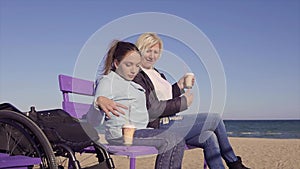 Young Pretty disabled woman in wheelchair sits on a banch near the sand sea beach and drinking coffee with her mother