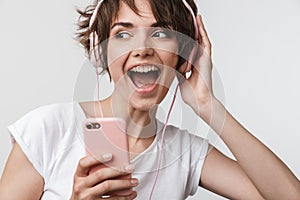 Young pretty excited happy woman posing isolated over white wall background using mobile phone listening music with earphones