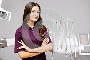 A young pretty female dentist is standing by the dental chair in the office with her arms crossed in front of her