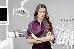 A young pretty female dentist is standing by the dental chair in the office with her arms crossed in front of her