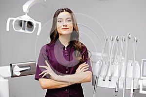 A young pretty female dentist is standing by the dental chair in the office with her arms crossed in front of her