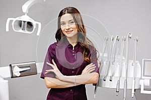 A young pretty female dentist is standing by the dental chair in the office with her arms crossed in front of her