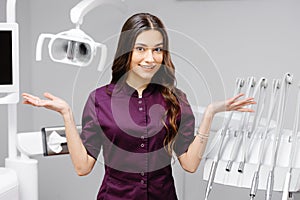 A young pretty female dentist is standing near the dental chair in the office, gesturing with her hands