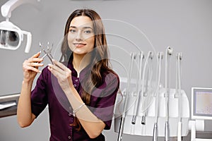 A young pretty female dentist is standing near the dental chair in the office, holding tools for work and looking into the camera