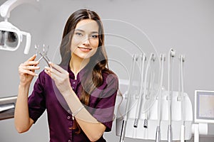 A young pretty female dentist is standing near the dental chair in the office, holding tools for work and looking into the camera
