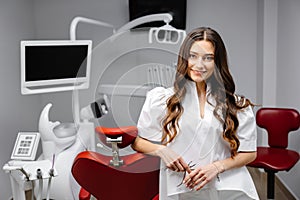 A young pretty female dentist is standing near the dental chair in the office, holding tools for work and looking into the camera