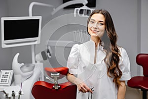 A young pretty female dentist is standing near the dental chair in the office, holding tools for work and looking into the camera