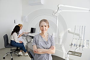 A young pretty female dentist stands in the middle of the office, and in the background a colleague is advising patients