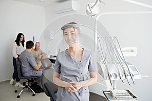A young pretty female dentist stands in the middle of the office, and in the background a colleague is advising patients