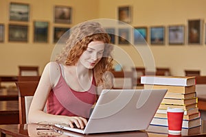 Young pretty female student with laptop and books working in high school library, sitting at table, takeaway cooffe near her books