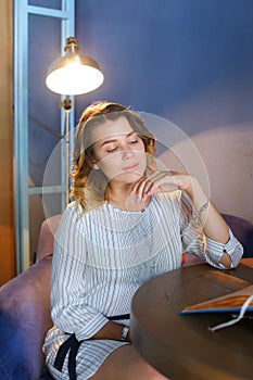 Young pretty female woman looks away with downcast eyes and smiles, holds hand near face and sits in chair at table on