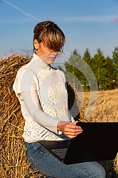 Pretty girl on a background of blue sky in a field near a haystack in a white jacket works at a laptop