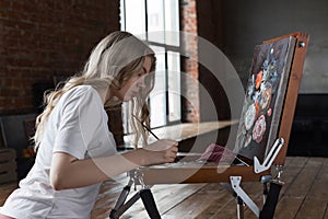 Young pretty girl with brush and palette sitting near easel drawing picture. Art, creativity, hobby, drawing process