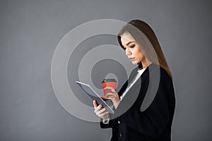Young pretty girl in business style clothes stands on a gray background with a tablet and a cup of coffee in her hands. Business