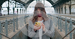 Young pretty girl in cotton handmade face mask looking and touching phone screen. Crop view of millennial brunette