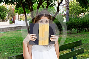 Young pretty girl is covering her face with a book because of feeling surprise and shock. Emotional funny woman is holding a book