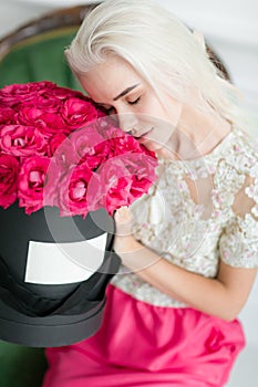 Young pretty girl with cute face and long blond hair. Woman sits and holding black box with pink roses. Luxury interior