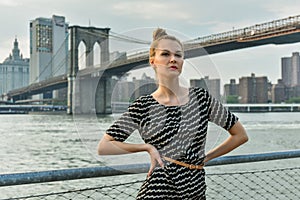 Young pretty girl elegantly dressed posing on the pier with Brooklyn Bridge on the background.