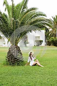 A young pretty girl of European appearance in a hat stands on a green lawn under palm trees. Tropical island. Concept of