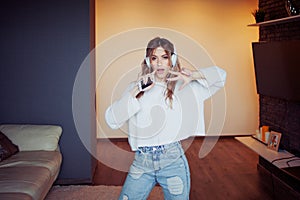 Young pretty girl in headphones with the phone. Listening to music, dancing and singing, indoors