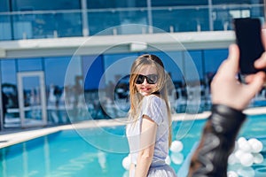 Young pretty girl with long hair and in black sunglasses is standing near pool. She wears gray T-shirt. Somebody hand