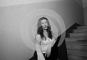 A young pretty girl with loose hair in a t-shirt and trousers is sitting in the entrance of the house on the landing with a seriou