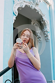 A young pretty girl in a purple dress paints her lips with lipstick, near the blue facade of a beautiful old building