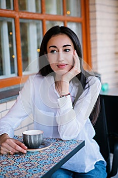 A young pretty girl is sitting at a table in a cafe and drinking coffee or tea