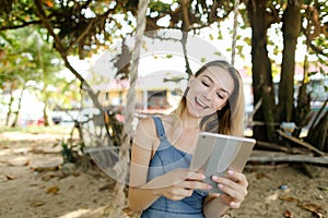 Young pretty girl using tablet and riding swing on sand, wearing jeans sundress.