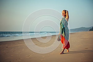 Young pretty girl walking along the seashore and admires the sunset. Blonde in bright clothes on sandy beach in the light of