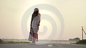 A young, pretty girl is walking in a long dress on the road barefoot in a summer, warm evening