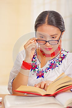 Young pretty girl wearing white shirt with colorful flower decorations and glasses, sitting by desk reading book smiling