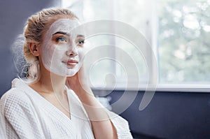 Young pretty girl with a white moisturizing mask applied to her face. A blue eyed blonde in a white robe does spa treatments in a